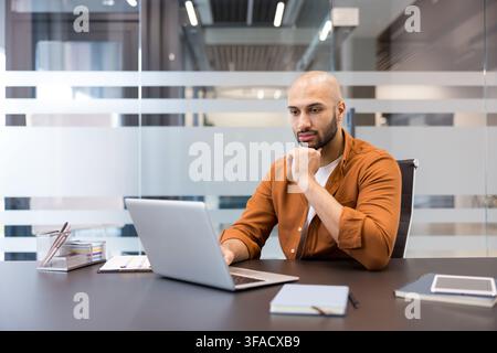 Young man concentrating on screen, working remotely or studying online, contemplating project details while using computer in a contemporary workspace Stock Photo