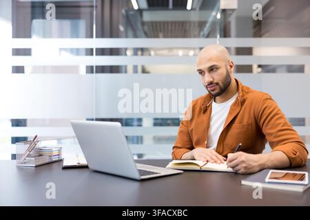 Young man focused on working, sitting at a modern office desk with a laptop, taking notes in a planner, representing business, learning, and productivity concepts Stock Photo