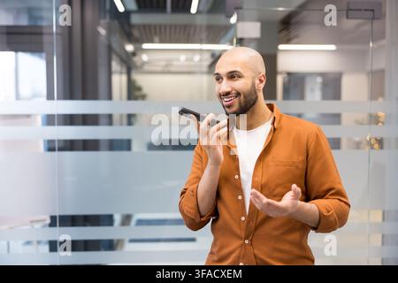 Young bald man smiling and talking into smartphone, recording a voice message or using voice assistant in a modern office environment Stock Photo