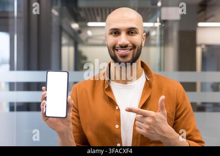 Happy young man showing blank smartphone screen, presenting new application or product while standing in a modern office with glass partitions Stock Photo