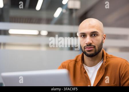 Young bald man with a beard looking directly at the camera while working on a laptop in a modern office environment, portraying focus and dedication Stock Photo