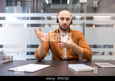 Young man wearing a headset and an orange shirt actively explaining a concept during a video call, offering expert advice and technical assistance from his office desk Stock Photo