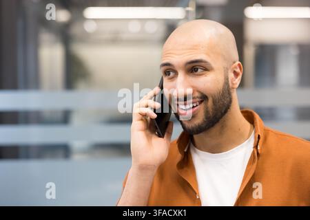 Smiling bald man with a beard actively engaging in a conversation on his mobile phone, communicating effectively in a bright office environment, expressing happiness and connectivity Stock Photo