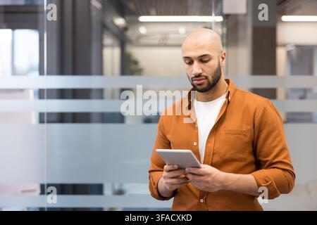 Young bald man with a beard concentrating on a digital tablet in his hands, standing casually in a contemporary office building with blurred glass partitions in the background Stock Photo