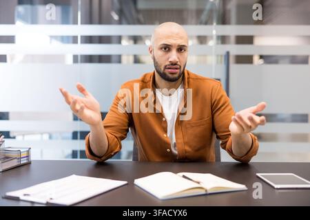 Young businessman sitting at office desk gesturing with hands, questioning or expressing confusion and uncertainty in a professional corporate environment Stock Photo