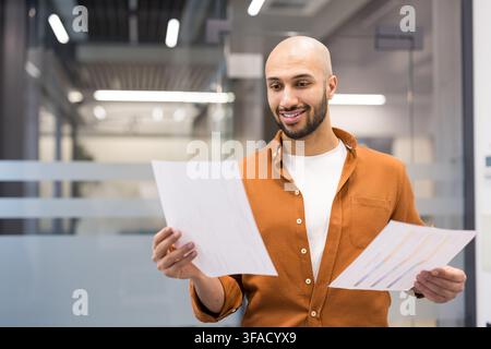 Happy professional man reviewing financial reports and paper documents, achieving success in his business career while working in a modern office environment Stock Photo