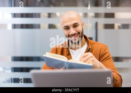 Young man smiling while making notes in a notebook, working happily at his laptop in an office, embodying productivity and a positive professional attitude Stock Photo