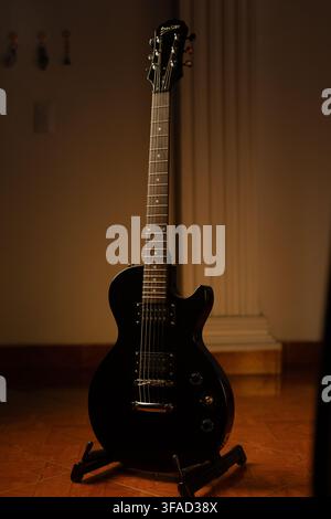 Vertical full length shot of a black solid-body electric guitar resting on a stand, showing its headstock, fretboard, humbucker pickups, and glossy bo Stock Photo