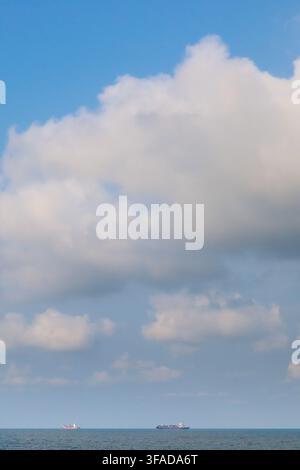 Dramatic white cumulus clouds over a calm blue ocean horizon with two distant cargo ships, Gulf of Thailand, vertical seascape. Stock Photo