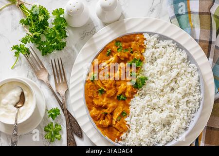 Spiced chicken fillet curry with cooked long grain rice, served on a plate with forks, yougurt, napkin on light marble backdrop, horizontal view, flat Stock Photo