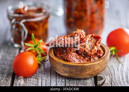 Bright sun-dried tomatoes marinated in olive oil served in a wooden bowl with fresh vine tomatoes and glass jars on a rustic wooden table, evoking Med Stock Photo