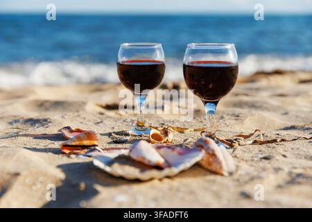 Two glasses of red wine on sandy beach with seashells and ocean waves relaxed summer coastal lifestyle romantic seaside mood Stock Photo