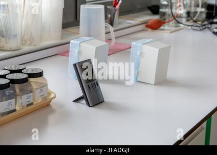 Rows of labeled sample bottles containing water on tray in laboratory, alongside a digital indoor-outdoor thermometer displaying temperature readings Stock Photo