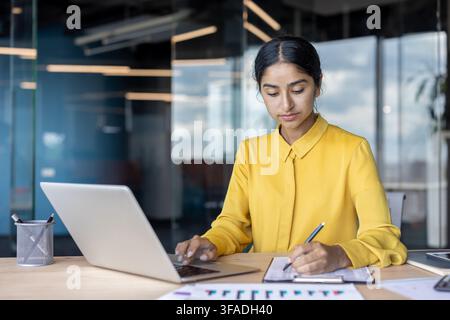 A young Indian businesswoman is busy taking notes in a document and typing on a laptop in the office. Stock Photo