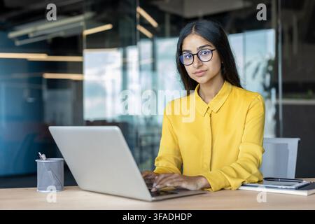 Confident young Indian businesswoman wearing glasses sitting at office desk, working in office, looking at camera. Stock Photo