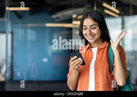 Happy young Indian woman standing in the office, holding a backpack, looking at the phone screen and rejoicing. Stock Photo
