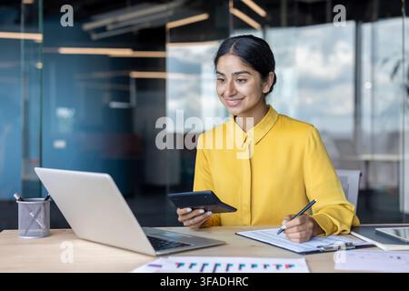 Smiling young Indian businesswoman working in the office at a desk with a laptop, taking notes and using a calculator. Stock Photo