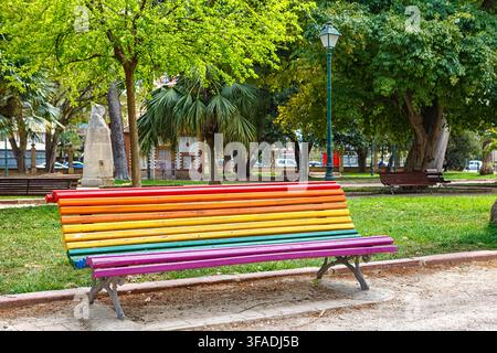 A vibrant bench painted in rainbow colors sits in a lively park filled with lush trees and grass. Stock Photo