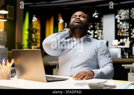 African american guy does self massage to relieve neck pain and stiffness, dealing with tension due to incorrect office posture and workload late at night. Stretching his upper back. Stock Photo