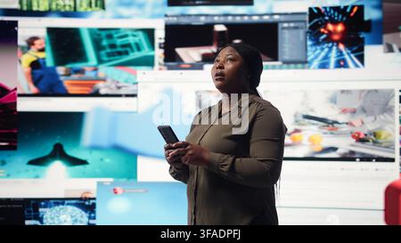 Woman browsing social media feed on smartphone in front of digital video wall. Person using mobile phone, floating icons highlight interactive engagement online, camera A Stock Photo