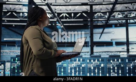 Admin in data center using app on laptop to ensure error free rigs networks for intense computational demands. IT expert uses notebook to monitor server hub gear components for failures, camera A Stock Photo