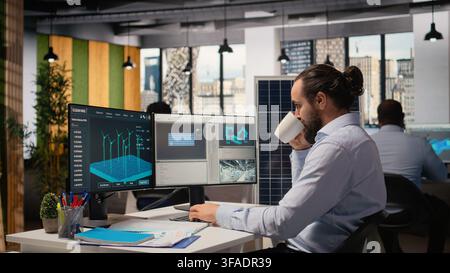 RD specialist develops clean energy solutions, drinking coffee, talking with coworker. Engineer chatting with colleague, enjoying beverage, optimizing renewable power generation on PC, camera A Stock Photo