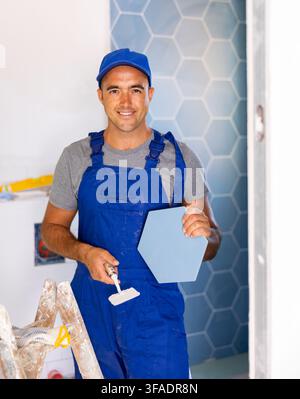 Skilled builder in work clothes posing with ceramic tiles and construction tools while repair and decoration in apartment Stock Photo