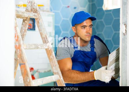 Focused construction worker in work clothes sitting squat engaged in wall tiling work during overhauls Stock Photo