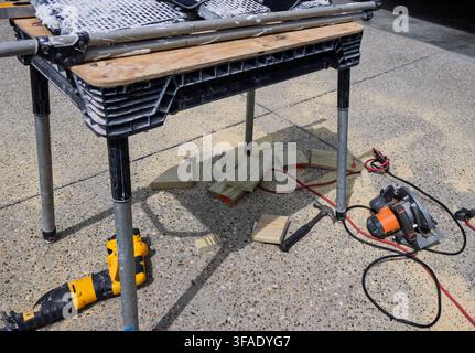 Folding workman's table with pipe legs and wooden planks. Metal legs stand on concrete ground. Yellow drill rests nearby. Circular saw lies on the pav Stock Photo