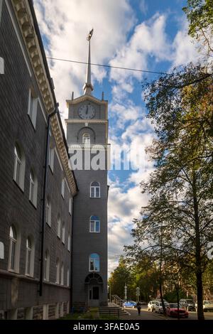 Petrozavodsk, Karelia, Russia, 15 August 2026 Tall narrow gray clock tower with spire and weathervane beside building, trees, street, cloudy sky. the Stock Photo