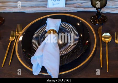 Elegant black table setting with gold accents, textured dinnerware, crystal glassware, and refined gold flatware on a dark wooden table Stock Photo