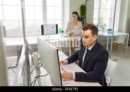Businessman working at desktop as businesswoman reviews documents in bright office Stock Photo
