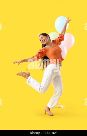 Celebration and joy, happy young woman with party hat and balloons dancing on a vibrant yellow background Stock Photo