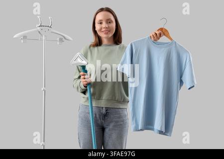 Home laundry and garment care, woman steaming a blue t-shirt with a clothes steamer on a stand, isolated on gray background Stock Photo