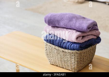 Wicker basket with stack of knitted sweaters on bedside bench in bedroom, closeup Stock Photo