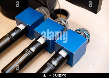 Three microphones sit side by side on a light wood surface. Blue cube-shaped covers protect their tops. The mics appear ready for use at an event. Ide Stock Photo