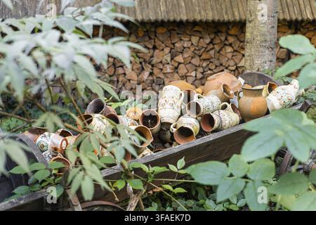A collection of traditional clay pots in various sizes and designs, resting on a wooden cart surrounded by green foliage and stacked firewood in the b Stock Photo
