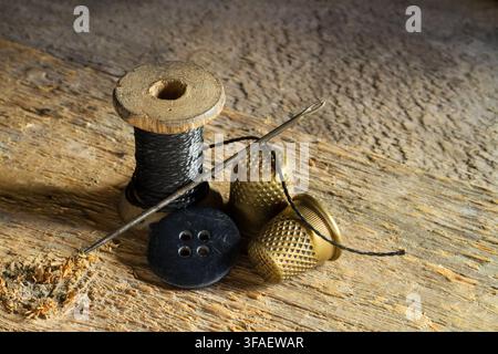 Antique sewing accessories. Tailor's accessories against the background of rough wood. Wooden spool of thread, copper thimbles, needle, button. Stock Photo