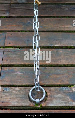 A bright steel chain runs vertically across weathered wooden decking, secured to a round mooring ring, with moss between the planks for a rustic water Stock Photo
