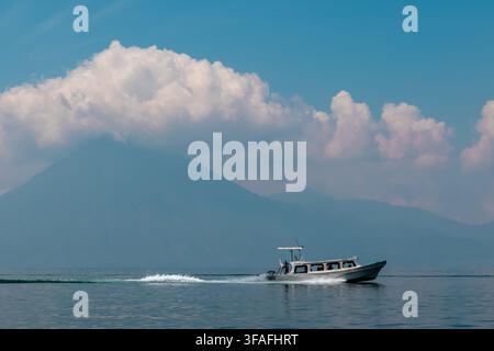 Small motorboat crossing calm blue water with a large mountain and soft white clouds in the background, creating a peaceful travel and nature scene. Stock Photo