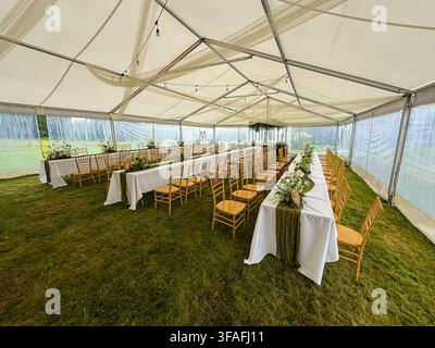 Riga, Latvia - August 1, 2026: Elegant outdoor wedding reception setup inside a spacious event tent featuring long banquet tables, wooden chairs, flor Stock Photo