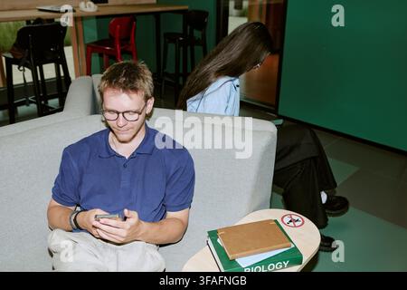 Diverse Students Using Smartphones on Sofa in University Campus Lounge Stock Photo