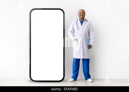 Full-length mature Black doctor in white coat standing beside oversized blank smartphone mockup on white studio background, ideal for healthcare app a Stock Photo