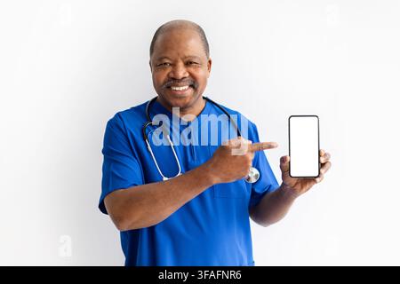 Smiling mature Black doctor in blue scrubs pointing to a blank smartphone screen against white studio background, ideal for healthcare apps, telemedic Stock Photo