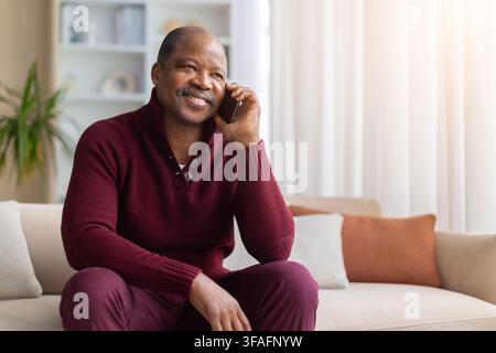 Smiling mature Black man sitting on sofa at home talking on smartphone Stock Photo