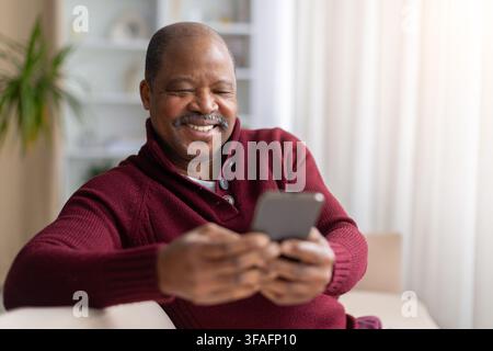 Smiling mature Black man relaxing on sofa at home while using smartphone Stock Photo
