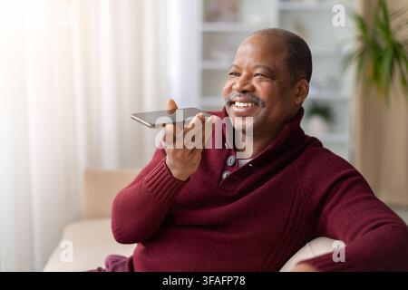Cheerful mature black man using voice assistant on smartphone at home Stock Photo