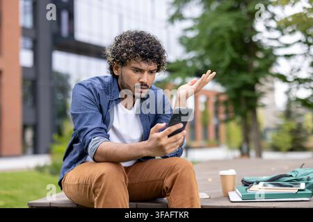 Young man sitting on a bench outdoors, looking confused and frustrated at his mobile phone, gesturing with his hand while having communication issues Stock Photo