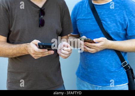 Two Men Using Smartphones Side by Side. Two men standing side by side using smartphones while dressed casually against a light neutral background. Stock Photo