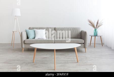 White table, modern sofa with white and blue pillows, lamp and vase with dry plants at home with gray walls Stock Photo